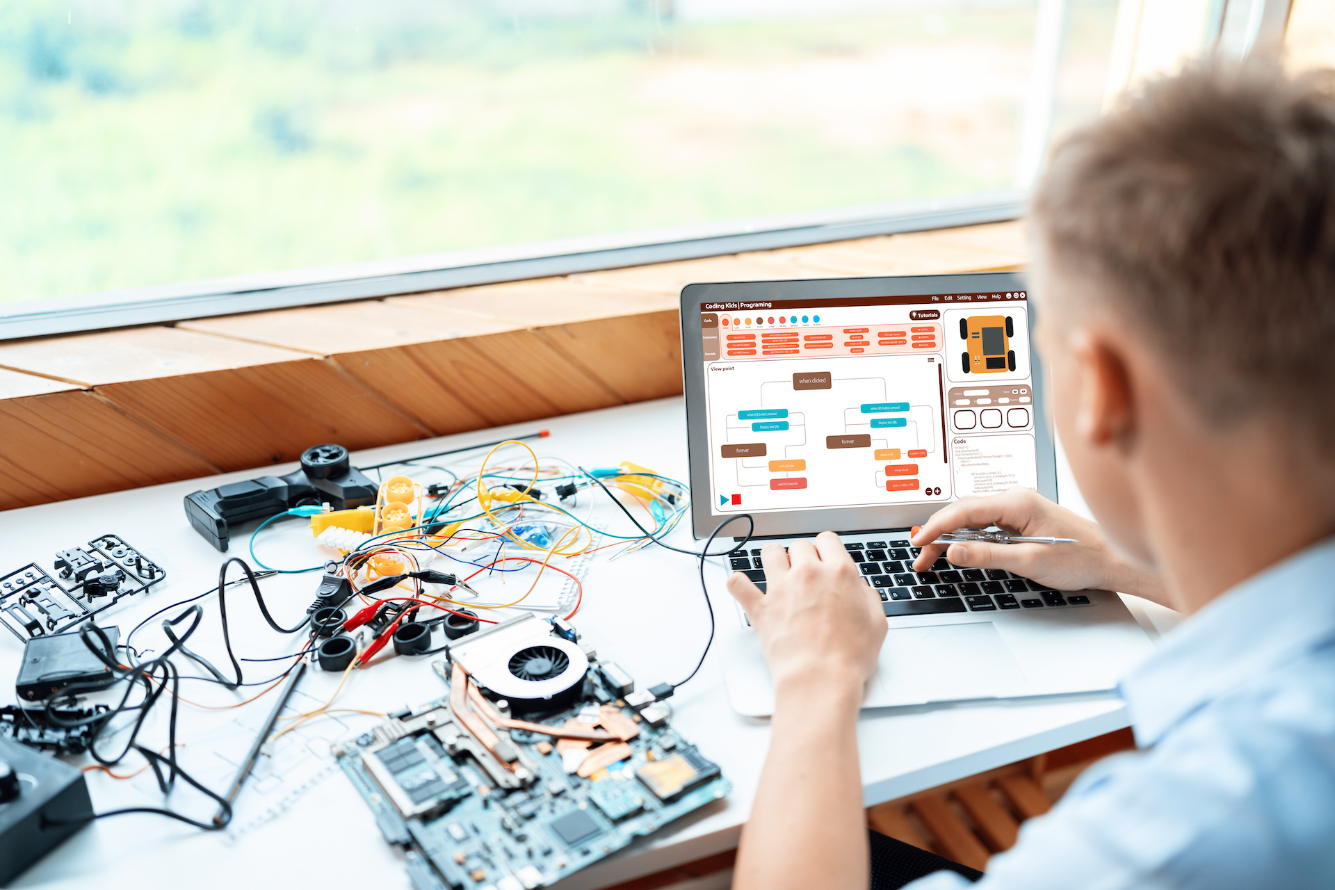 Blonde hair schoolboy in blue shirt code robotics car use laptop learn technology and engineering in STEM class. Around laptop put controller, electric wire, battery charger and laptop. Edification.